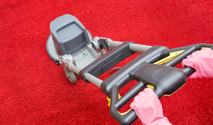 Pink-gloved person using a vacuum to clean and remove stains from a red carpet.