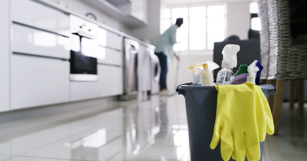 Cleaning supplies bucket with spray bottles and yellow gloves in a modern white kitchen.