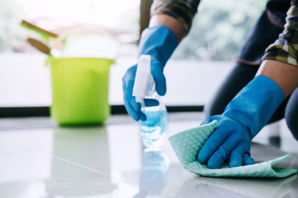 Person wearing blue gloves spraying cleaner and wiping a tile floor with a cloth.