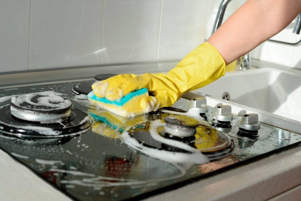 A person in yellow gloves scrubs a stove top focusing on removing stains and ensuring cleanliness