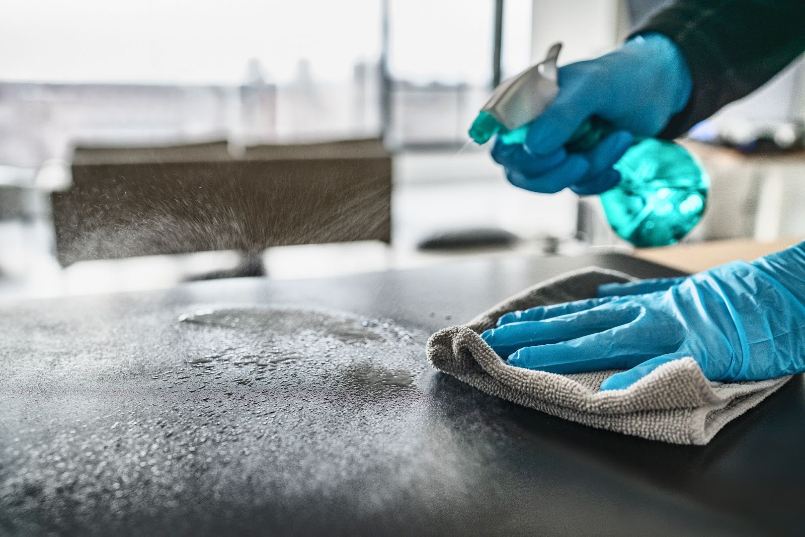 A person in blue gloves sprays cleaner on a surface, wiping it with a cloth in a modern room.