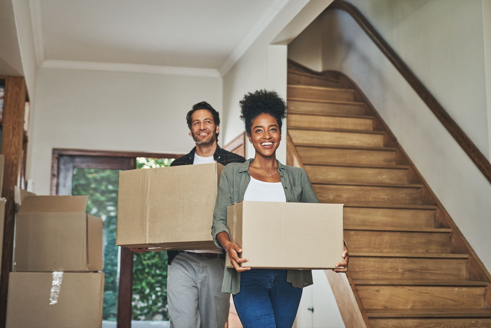 A smiling couple carrying moving boxes inside a house with a wooden staircase in the background.