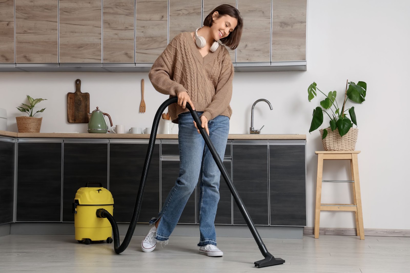 A smiling woman wearing a brown sweater and jeans vacuuming the kitchen floor with a yellow canister vacuum.