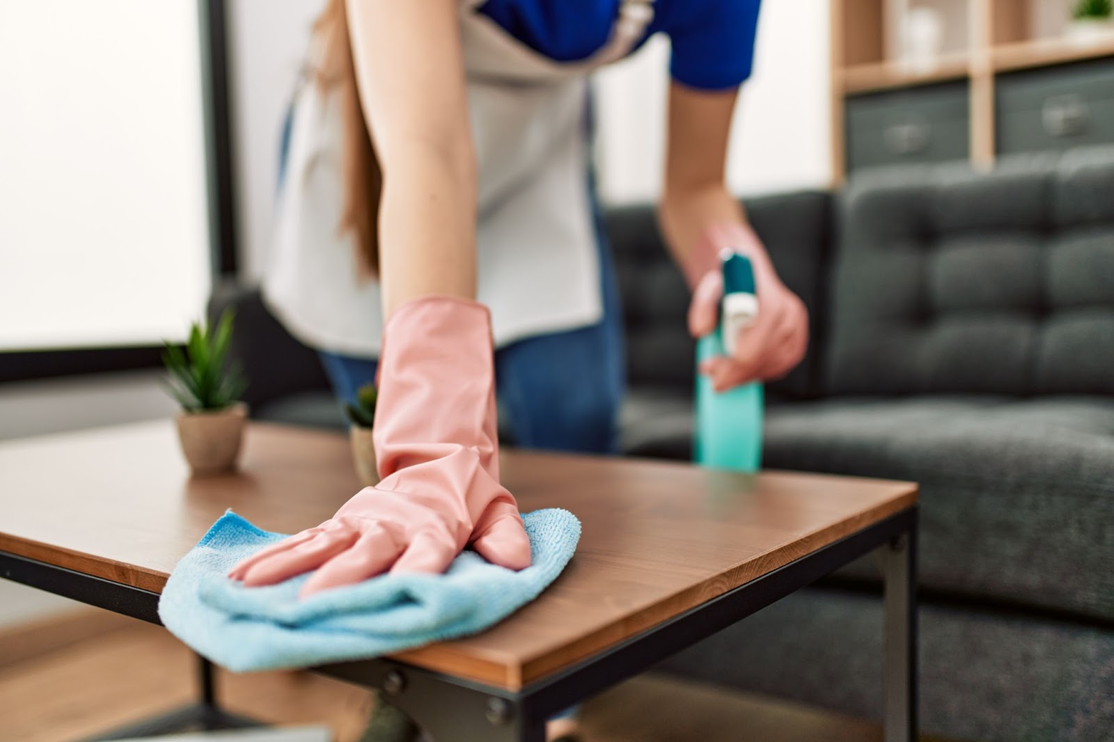 A person wearing pink rubber gloves and an apron cleans a wooden coffee table with a blue microfiber cloth while holding a spray bottle.