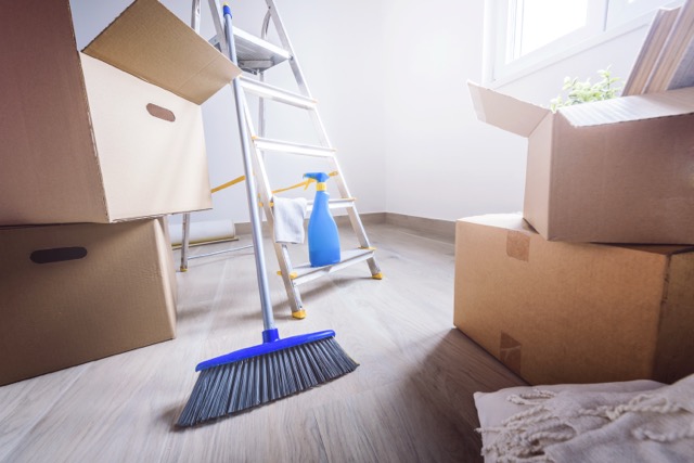 A blue broom, a spray bottle on a step ladder, and several cardboard boxes in a bright, empty room.