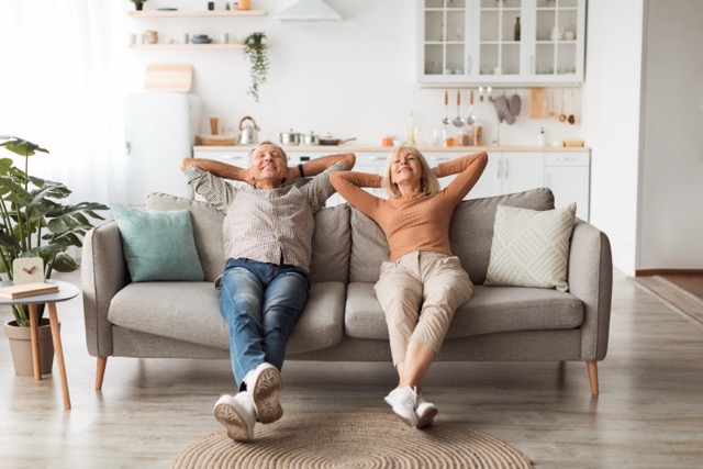 A happy senior couple relaxing on a grey sofa with their hands behind their heads in a bright, clean living room.