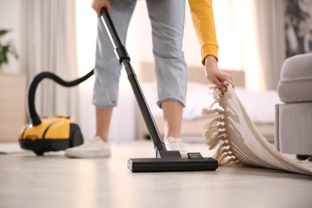 A person lifting the edge of a fringed rug while vacuuming a light-colored floor with a yellow canister vacuum.