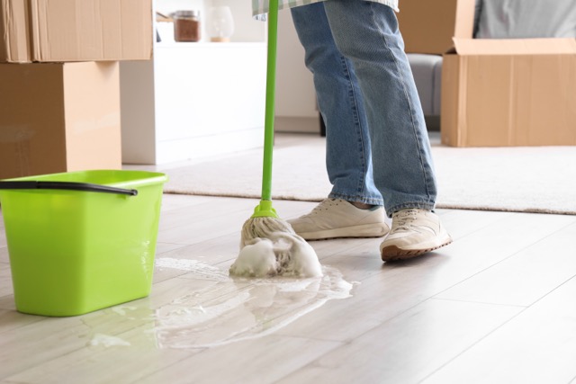 A person mopping a soapy spill on a light wood floor next to a bright green bucket and cardboard boxes.