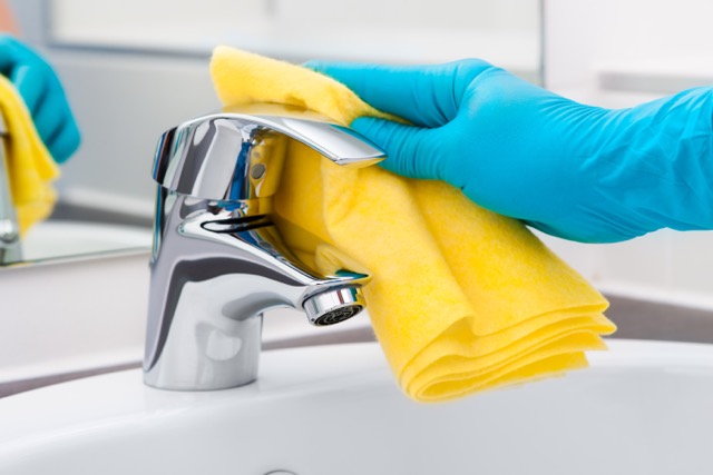 A person wearing blue protective gloves using a yellow cloth to polish a chrome bathroom faucet.