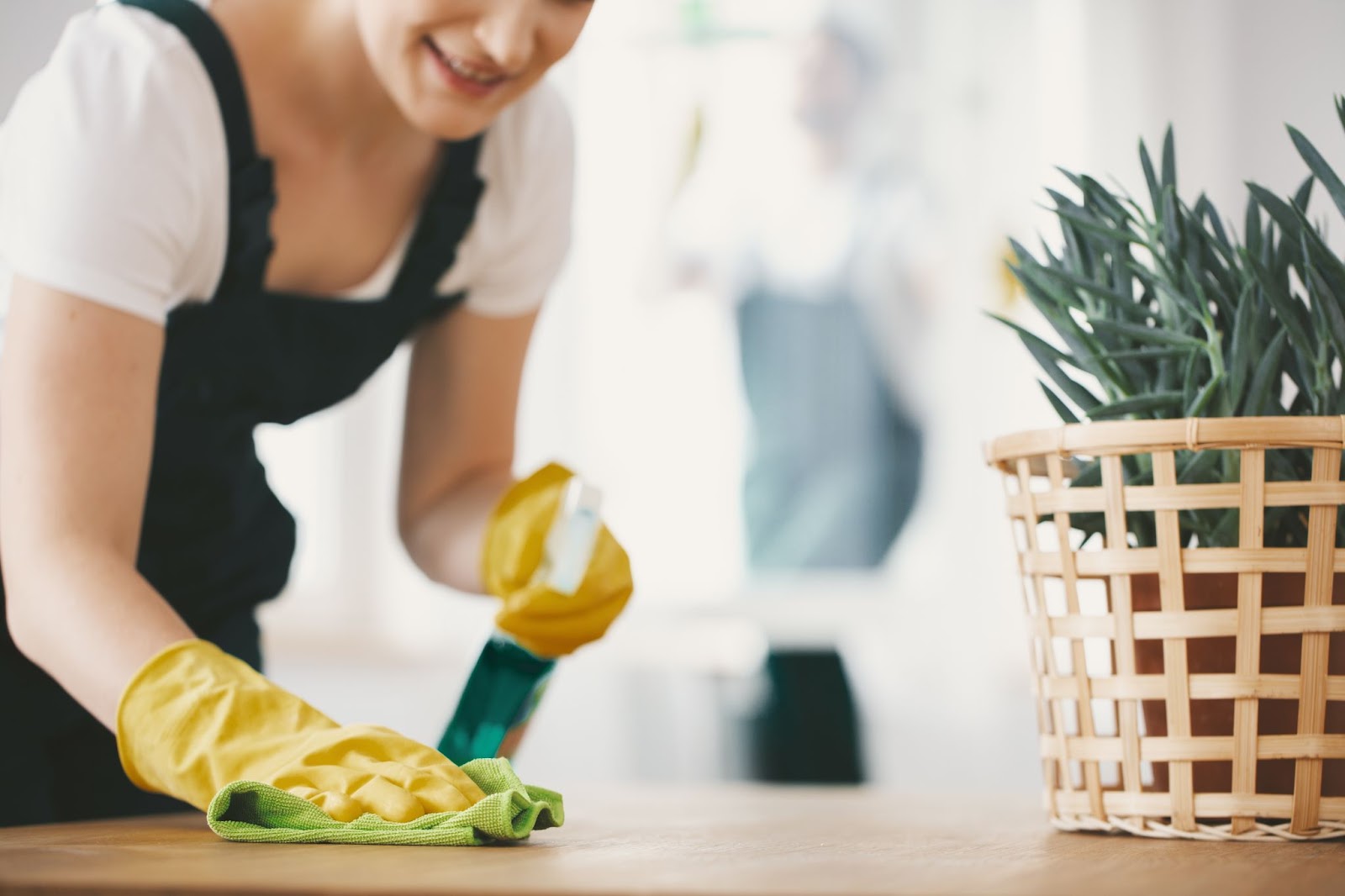 A smiling person wearing yellow rubber gloves and a black apron wiping a wooden surface with a green cloth.