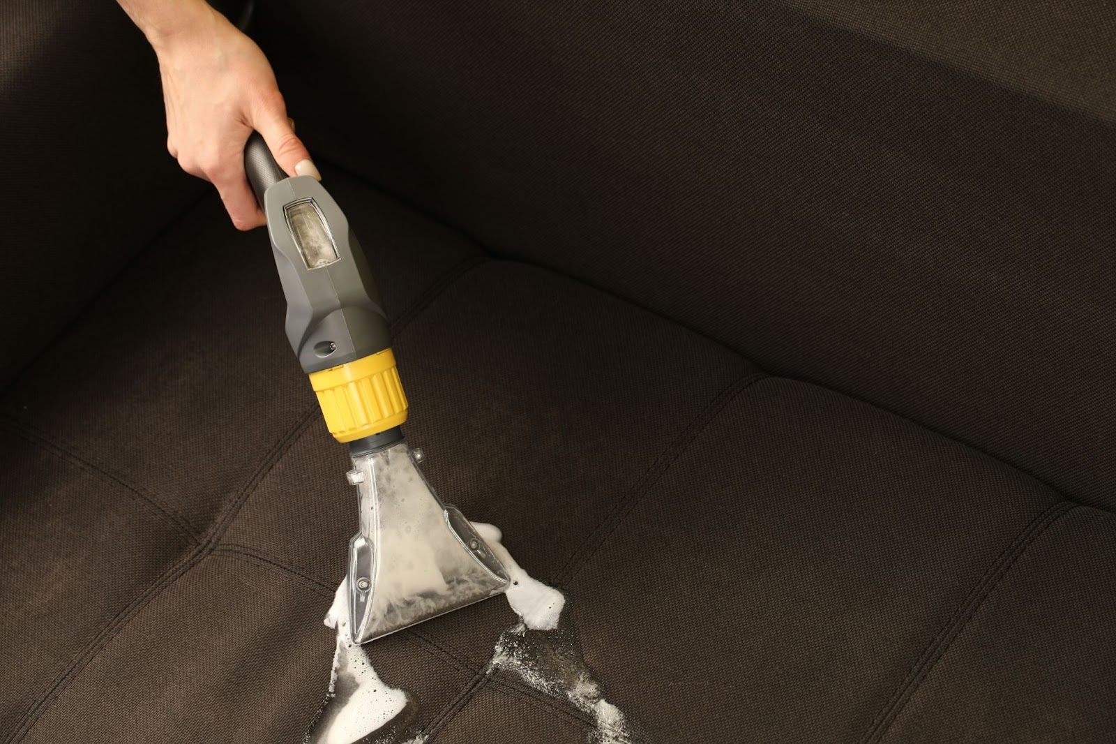 A person using a steam cleaner attachment and soapy foam to deep clean a dark fabric sofa.
