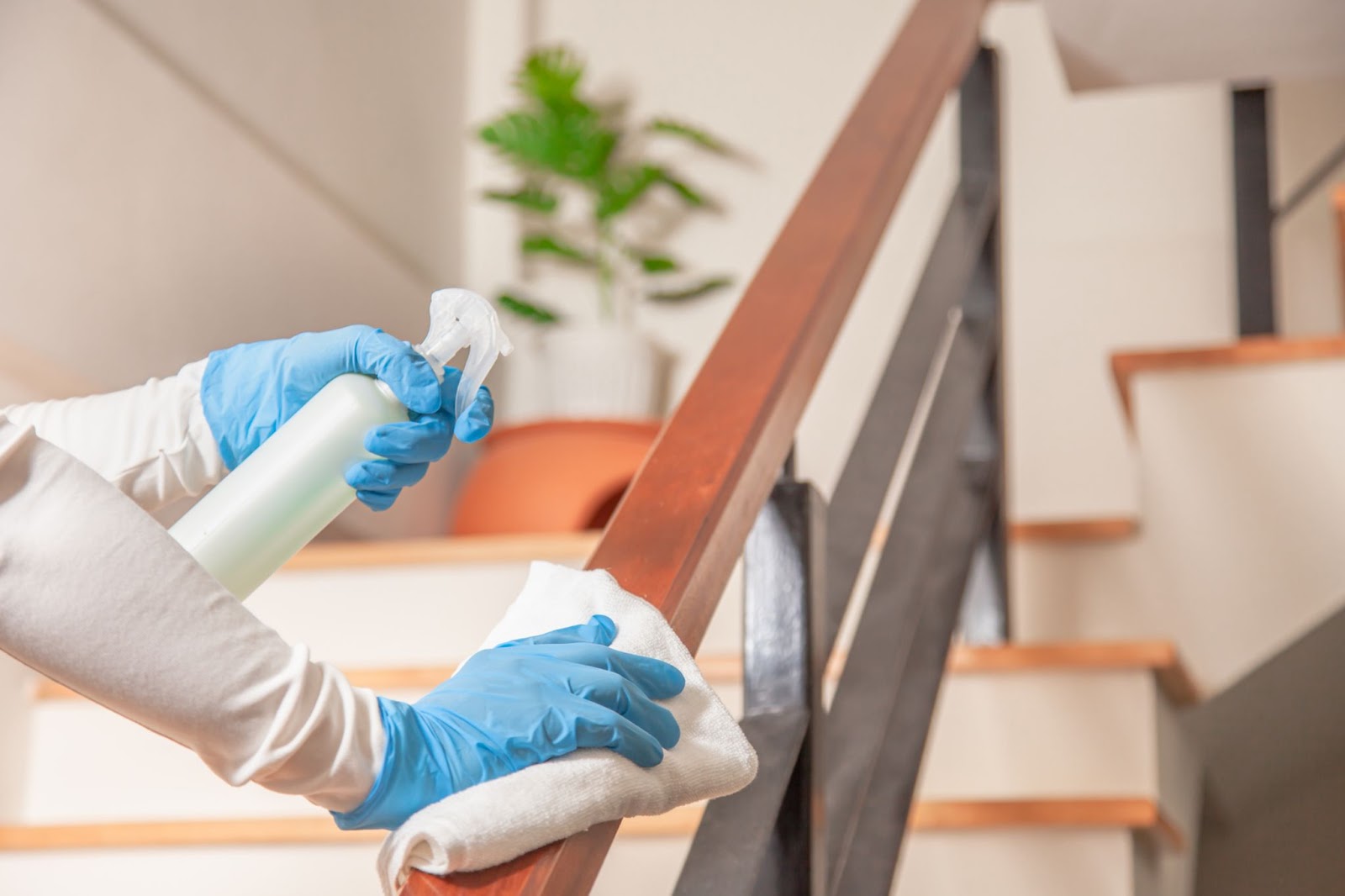 A person wearing blue gloves using a spray bottle and a white cloth to disinfect a wooden staircase railing.