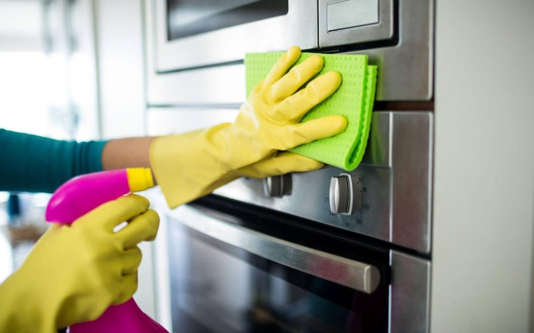 Hands in yellow gloves clean a stainless steel oven with a green cloth and pink spray bottle
