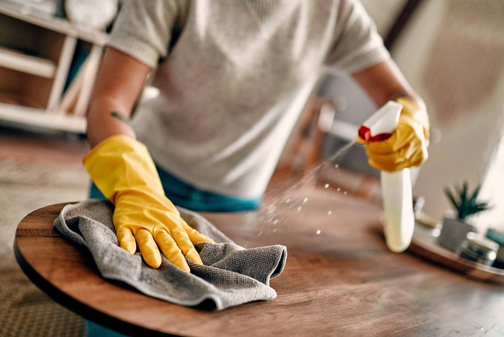 Person wearing yellow gloves cleaning a wooden table with a gray cloth and spray bottle