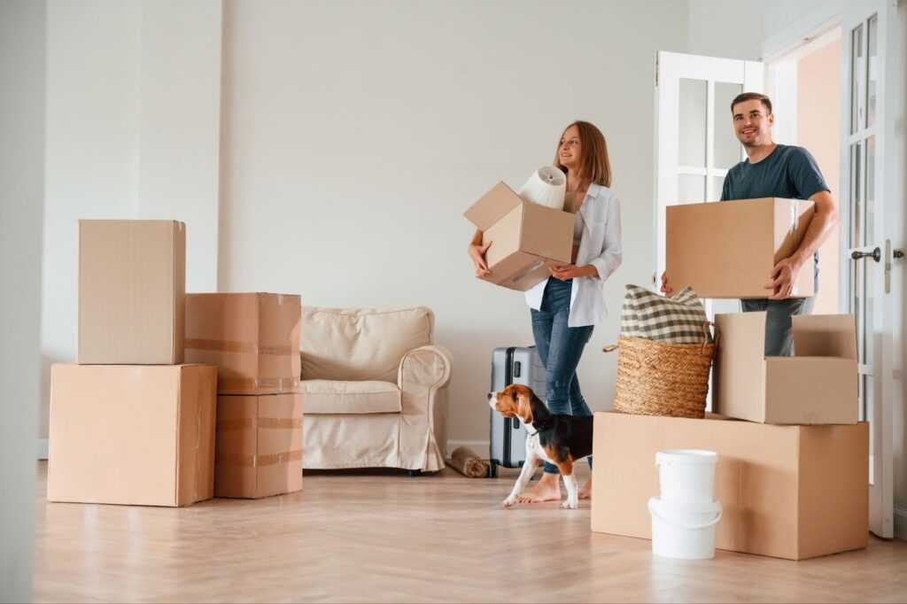 A couple moving into a new home with cardboard boxes, a beagle dog, and household items around a beige sofa.