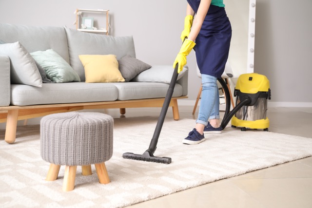 A person in an apron and rubber gloves using a yellow vacuum cleaner to clean a light-colored rug in a modern living room.