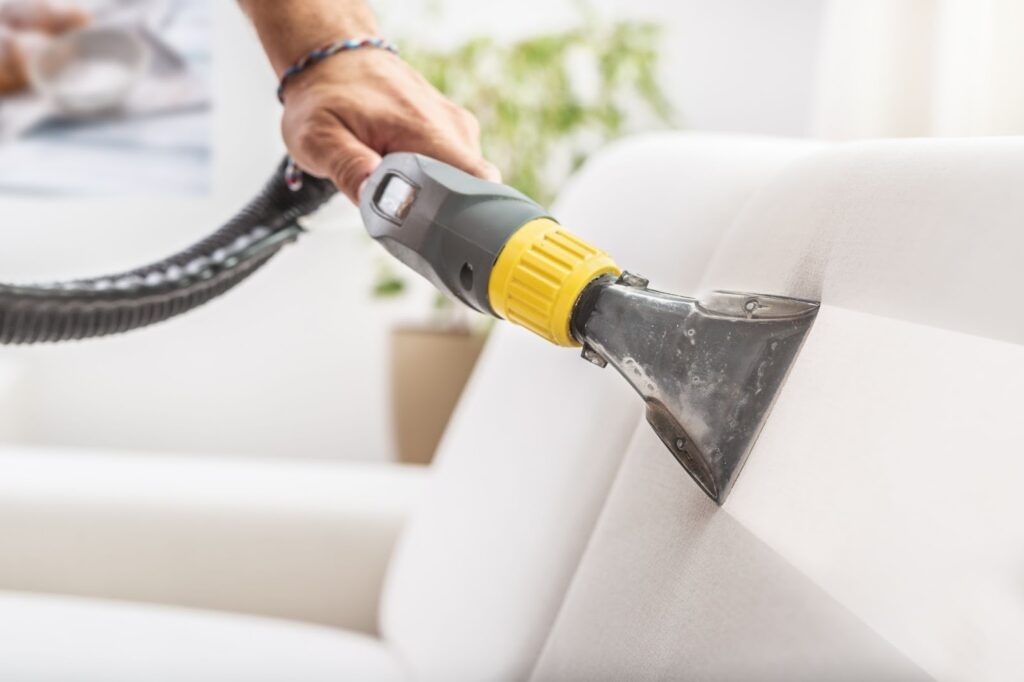 A person using a specialized vacuum attachment to deep clean a white fabric sofa.