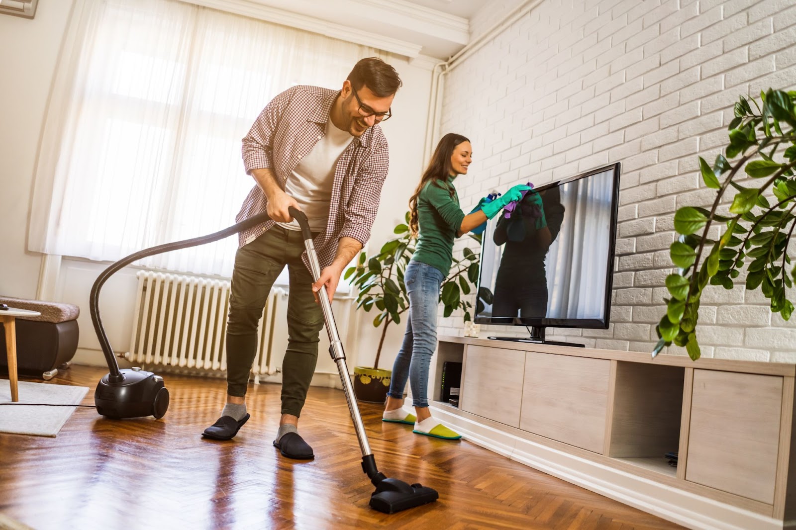 A smiling couple cleaning their living room together, with the man vacuuming the floor and the woman wiping down the television screen.