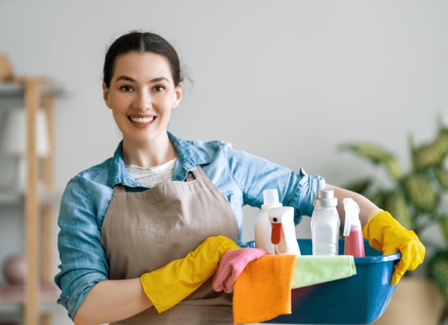 A smiling woman wearing yellow rubber gloves and an apron, carrying a blue basin filled with cleaning supplies and colorful cloths.