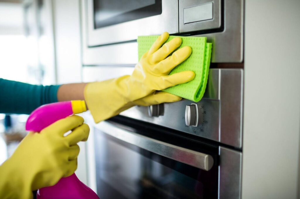 Hands in yellow gloves clean a stainless steel oven with a green cloth and pink spray bottle