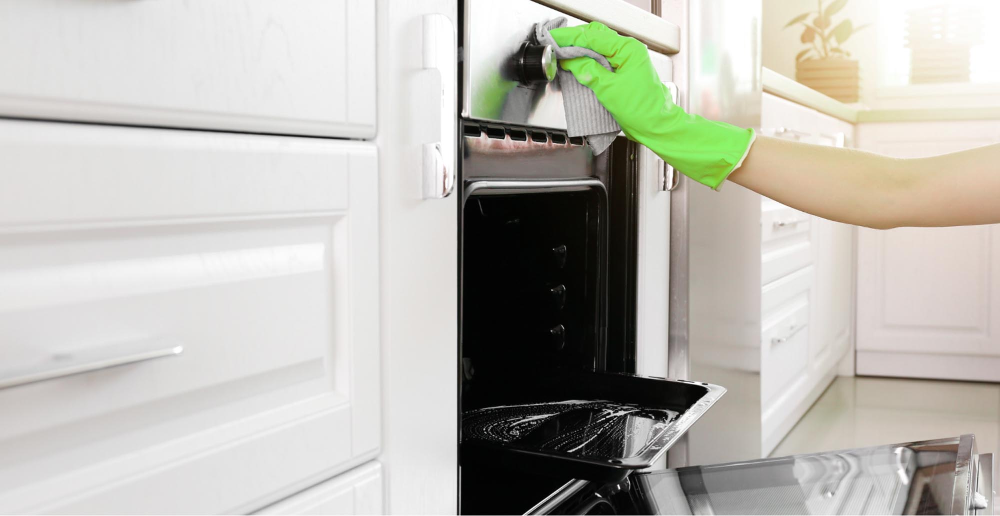 A woman wearing a white shirt and green gloves is cleaning an oven, demonstrating proper cleaning techniques.