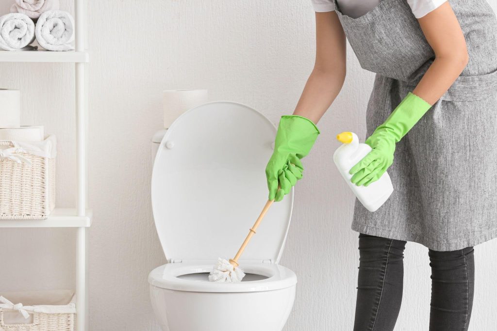A woman wearing a green glove is cleaning a toilet with a brush focused on maintaining cleanliness